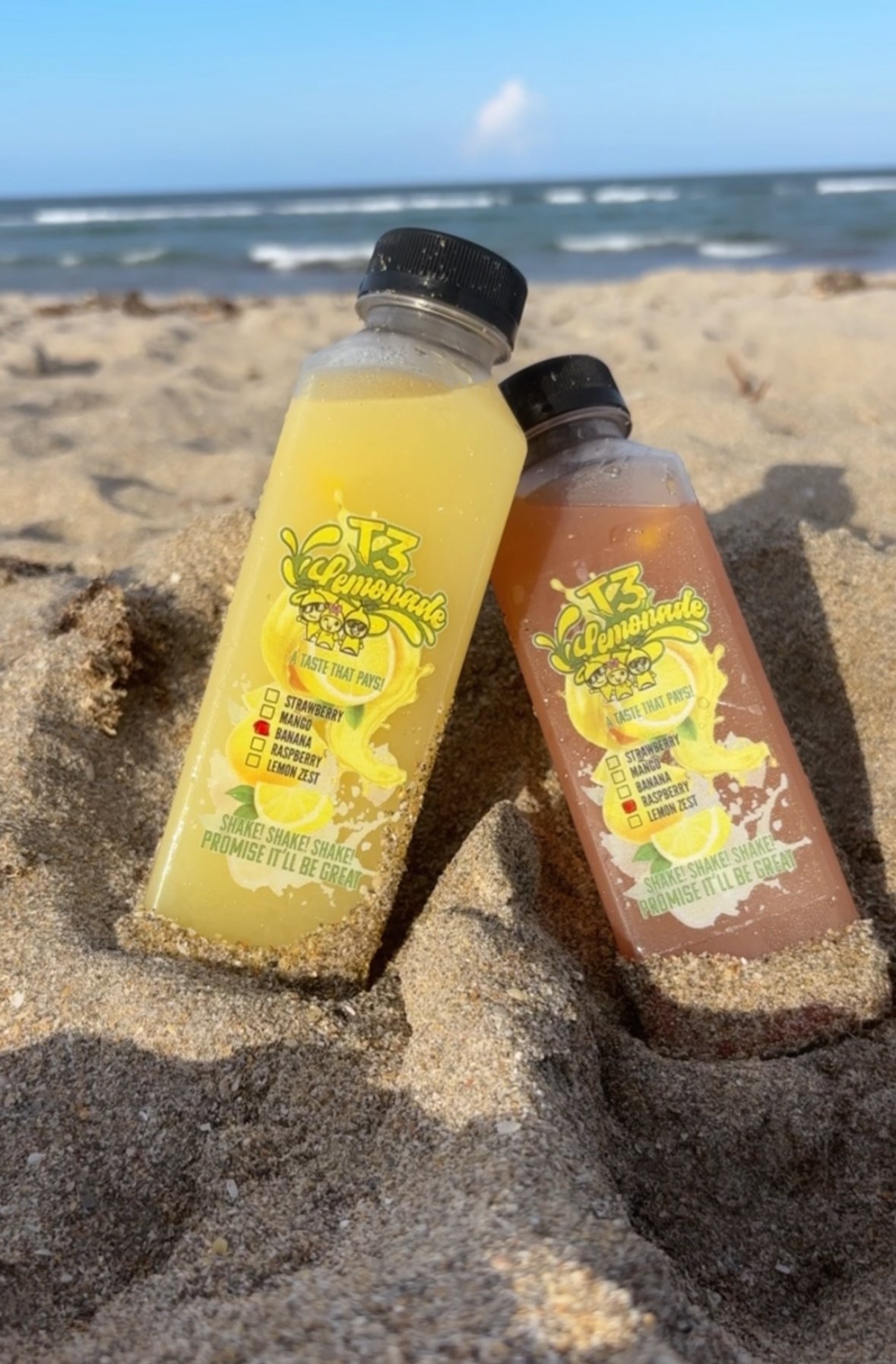 Two bottles of T3 Lemonade standing in beach sand